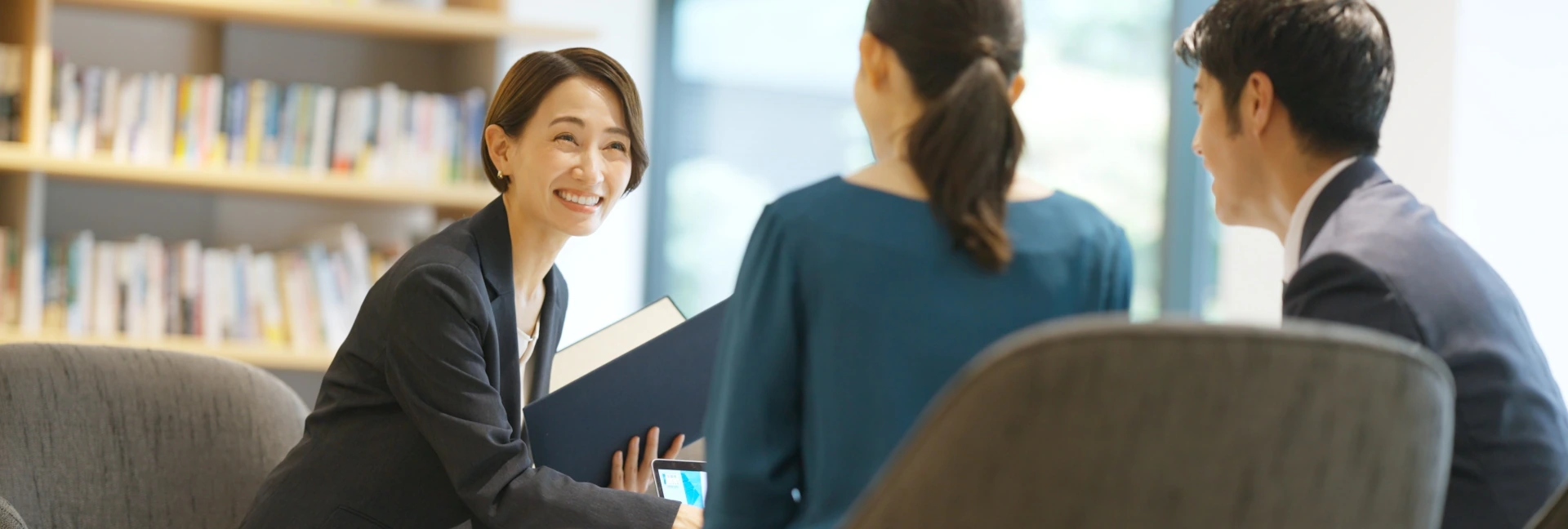 ノーマンジャパン採用ページ ショールームスタッフ募集 キービジュアル（PC用）　女性ビジネスウーマンが椅子に座り、二人のお客様と話している様子