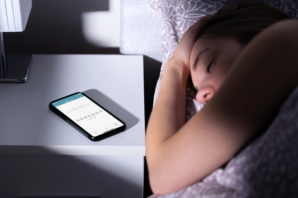 熟睡する女性の隣に、シェードのタイマー機能をセットしたスマートフォンがある写真